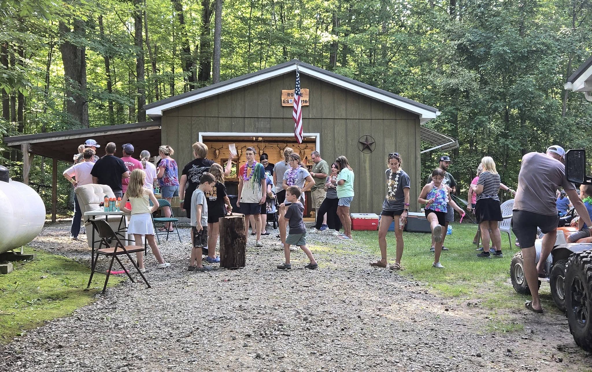 A crowd of neighbors, some wearing leis, gathered on the gravel drive of a pole-barn host house flying an American flag during the Sub Crawl.